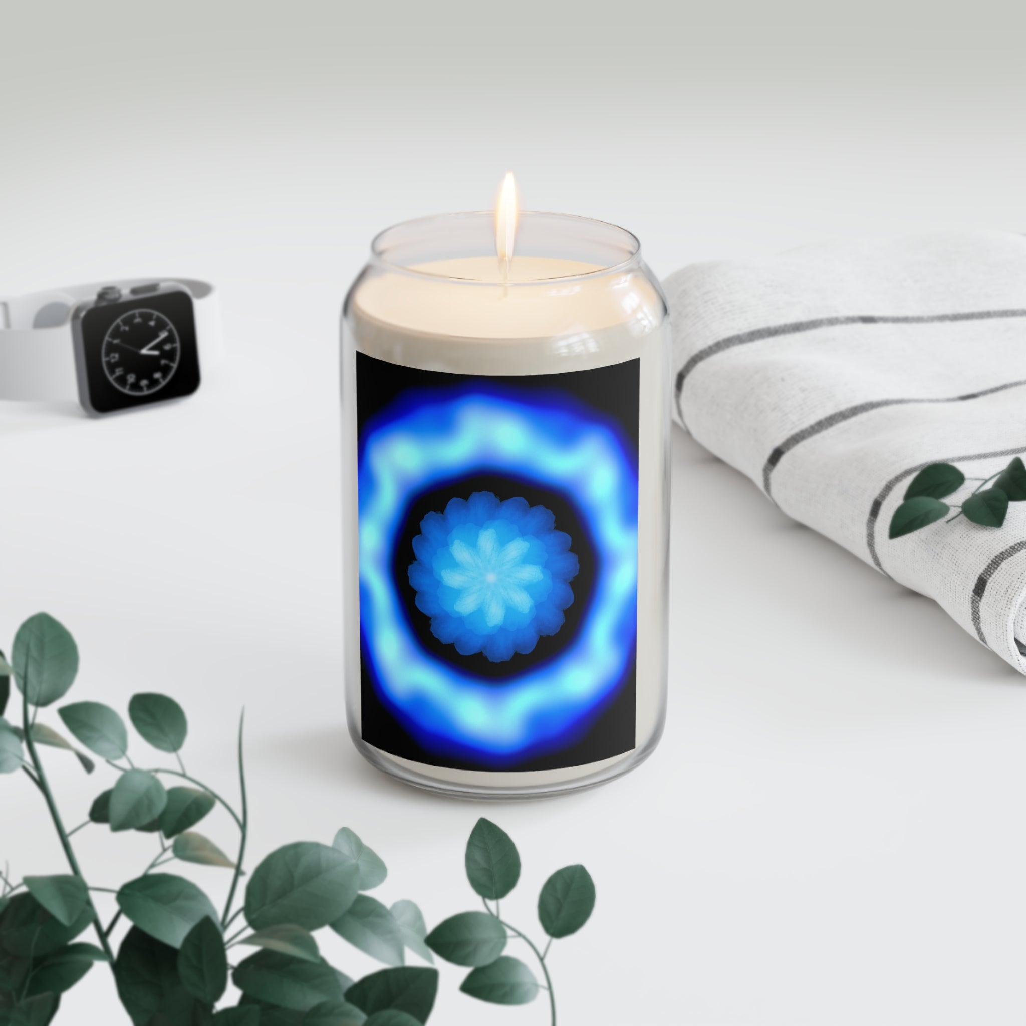 Candle in a glass jar with a blue and black design on a white surface with a plant and watch in the background.